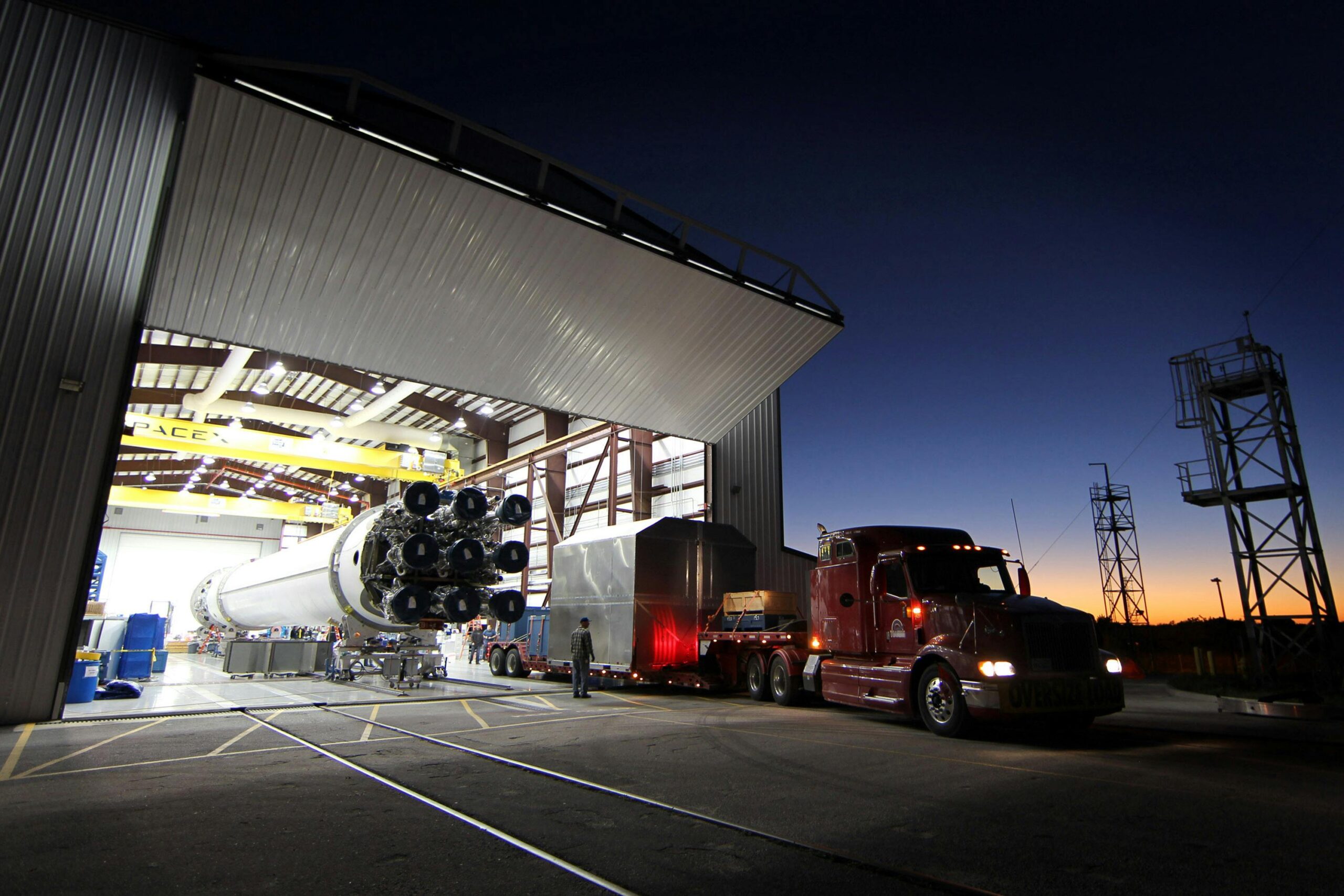 Modern cargo truck parked near hangar with massive rocket core inside at industrial factory in evening